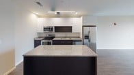 This thoughtfully designed single-floor unit offers a clean, contemporary aesthetic throughout. The open-concept kitchen serves as the centerpiece, featuring dark lower cabinetry contrasted by sleek white upper cabinets, granite countertops, a generous island for additional prep and seating space, and a full suite of stainless steel appliances including a refrigerator with ice maker, range, microwave, and dishwasher. Pendant and recessed lighting above the kitchen create a bright, functional workspace. Dark wood-grain luxury vinyl plank flooring flows through the main living areas and bedrooms, lending warmth and continuity to the space. The living area is spacious and airy, with a sliding glass door opening to a private balcony that offers expansive urban views. A large picture window in the bedroom further captures sweeping cityscape vistas, including notable architectural landmarks. Bedrooms are well-proportioned and include closet storage. Two bathrooms mirror each other in finish, each featuring granite-topped dark vanities, large-format light tile flooring, and a full tub-shower combination. Neutral wall tones throughout the unit complement the modern finishes, creating a cohesive and move-in-ready living environment.