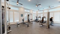 Located at 833 E Magnolia Dr in Tallahassee, Florida, this single-story fitness center offers a thoughtfully designed workout environment suited for a comprehensive exercise routine. The space is bathed in natural light, thanks to an array of large windows with white horizontal blinds lining multiple walls, complemented by transom-style windows positioned near the ceiling that draw in additional daylight and contribute to the room's open, airy feel. Multiple ceiling fans with integrated lighting are distributed throughout, ensuring consistent airflow and comfort during workouts.

The fitness center is equipped with a well-rounded selection of cardio machines, including treadmills, an elliptical trainer, and a recumbent stationary bike. Strength training is supported by a multi-function cable machine, adjustable weight benches, a dumbbell rack, and various resistance-based equipment. The durable speckled rubber flooring provides both safety and functionality underfoot.

A built-in dual water fountain station is conveniently mounted along one wall, offering easy hydration access. French doors provide direct outdoor access, adding a sense of connectivity to the surrounding environment. The neutral-toned walls and recessed lighting complete the clean, modern aesthetic of this practical and well-appointed fitness facility.