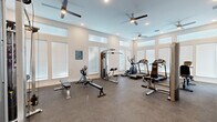 Located at 833 E Magnolia Dr in Tallahassee, Florida, this single-story fitness center offers a thoughtfully designed workout environment suited for a comprehensive exercise routine. The space is bathed in natural light, thanks to an array of large windows with white horizontal blinds lining multiple walls, complemented by transom-style windows positioned near the ceiling that draw in additional daylight and contribute to the room's open, airy feel. Multiple ceiling fans with integrated lighting are distributed throughout, ensuring consistent airflow and comfort during workouts.

The fitness center is equipped with a well-rounded selection of cardio machines, including treadmills, an elliptical trainer, and a recumbent stationary bike. Strength training is supported by a multi-function cable machine, adjustable weight benches, a dumbbell rack, and various resistance-based equipment. The durable speckled rubber flooring provides both safety and functionality underfoot.

A built-in dual water fountain station is conveniently mounted along one wall, offering easy hydration access. French doors provide direct outdoor access, adding a sense of connectivity to the surrounding environment. The neutral-toned walls and recessed lighting complete the clean, modern aesthetic of this practical and well-appointed fitness facility.