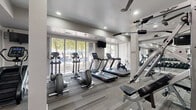 Located at 9680 W Northern Ave in Peoria, Arizona, this fitness center is a thoughtfully designed facility spread across a single floor. The space is organized into two interconnected exercise rooms, offering a comprehensive workout environment for a variety of training styles.

The main exercise area is bright and open, featuring large windows that invite natural light and provide views of the surrounding outdoor greenery. Patterned carpet tiles in neutral tones cover the floors throughout, lending a clean and modern aesthetic. Ceiling fans are strategically positioned overhead to maintain airflow and comfort during workouts. Recessed lighting further enhances the well-lit atmosphere.

Cardio equipment is prominently featured, including multiple treadmills, elliptical trainers, stationary bikes, and a stair climber. A dedicated free weight section houses a full rack of dumbbells, while strength training machines and cable systems offer options for resistance workouts. Wall-mounted mirrors expand the sense of space and allow users to monitor their form. A flat-screen TV is mounted for entertainment during workouts.

The secondary room serves as a transitional and supplemental workout space, with motivational wall graphics, informational fitness posters, and access to additional equipment. The entry area includes a sanitization station and clear signage for convenience. The overall ambiance is clean, modern, and purpose-driven, making it a fully functional fitness destination.