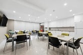 Located at 350 Elliott St in Windsor, Ontario, this communal lounge and kitchen space offers a bright, functional, and welcoming environment across a single floor. The open layout accommodates both dining and kitchen activities seamlessly, making it well-suited for shared use.

The dining area features multiple white rectangular tables surrounded by a mix of black and yellow-green upholstered chairs, providing ample seating for groups. Recessed ceiling lighting illuminates the space evenly, while roller blinds on the windows offer adjustable natural light. A wall-mounted flat-screen TV adds a social element to the lounge setting. Nature-themed artwork on the neutral beige walls contributes to a calm and inviting ambiance. Large-format white floor tiles run throughout, lending the space a clean, cohesive look.

The kitchen area is outfitted with an extensive run of white shaker-style cabinetry — both upper and lower — paired with neutral stone-look countertops. A double stainless steel sink, dishwasher, refrigerator, microwave, and coffee station ensure full functionality for communal use. Recessed lighting keeps the kitchen bright and practical. A labeled "Lounge" entrance door, fire safety equipment, and an electrical panel are also visible, reflecting the space's shared-facility character.