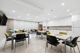 Located at 350 Elliott St in Windsor, Ontario, this communal lounge and kitchen space offers a bright, functional, and welcoming environment across a single floor. The open layout accommodates both dining and kitchen activities seamlessly, making it well-suited for shared use.

The dining area features multiple white rectangular tables surrounded by a mix of black and yellow-green upholstered chairs, providing ample seating for groups. Recessed ceiling lighting illuminates the space evenly, while roller blinds on the windows offer adjustable natural light. A wall-mounted flat-screen TV adds a social element to the lounge setting. Nature-themed artwork on the neutral beige walls contributes to a calm and inviting ambiance. Large-format white floor tiles run throughout, lending the space a clean, cohesive look.

The kitchen area is outfitted with an extensive run of white shaker-style cabinetry — both upper and lower — paired with neutral stone-look countertops. A double stainless steel sink, dishwasher, refrigerator, microwave, and coffee station ensure full functionality for communal use. Recessed lighting keeps the kitchen bright and practical. A labeled "Lounge" entrance door, fire safety equipment, and an electrical panel are also visible, reflecting the space's shared-facility character.