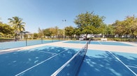 This outdoor recreational court facility offers multiple side-by-side pickleball and tennis courts finished in a striking two-tone blue and light gray surface coating. Each court is clearly lined and equipped with regulation nets, providing a polished and functional playing environment. The courts are fully enclosed by perimeter chain-link fencing, ensuring safety and defining the playing areas. Tall high-mast sports lighting poles are positioned throughout the facility, enabling play well into the evening hours.

Surrounding the courts, mature shade trees provide a natural canopy that softens the open-air setting, while benches placed along the sidelines offer comfortable seating for spectators and players at rest. The grounds are well-kept, with open green space visible beyond the fencing, contributing to an inviting park-like atmosphere.

The facility appears to be part of a larger community or residential campus, with buildings and landscaped areas visible in the background. Under clear, sunny skies, the bright blue court surfaces stand out vividly, lending the space an energetic and well-maintained character. Overall, this court complex is a well-appointed outdoor athletic amenity designed to support active recreational use for players of all skill levels.