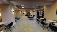This single-floor community space offers a thoughtfully designed environment that balances comfort, functionality, and a welcoming atmosphere. The open layout stretches generously from end to end, with recessed ceiling lighting and skylight panels casting a bright, even illumination throughout the carpeted interior. Patterned low-pile carpeting in neutral tones anchors the space and contributes to a calm, refined aesthetic.

The lounge area toward one end features plush teal upholstered seating, including a sofa and ottoman, ideal for quiet relaxation or casual conversation. Wall-mounted sconces line the corridor-style layout, adding a warm, ambient glow that complements the recessed lighting above. A wall-mounted flat-screen display serves as a focal point deeper in the space, flanked by additional seating arrangements.

The library section, situated prominently along one wall, houses tall dark wood bookshelves filled with a curated selection of reading material. Framed artwork adorns the surrounding walls, lending a cultured and inviting character to this area. Nearby, small round bistro tables paired with black chairs create intimate spots for reading or light conversation.

Overall, the space is versatile and well-appointed, seamlessly combining a social lounge, media area, and reading library into a cohesive and accessible community environment.