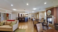 This single-floor community space offers an open, multipurpose layout designed to bring people together in a comfortable and functional environment. The lounge area at the front features a cushioned sofa and accent rugs, providing a relaxed seating arrangement for socializing or unwinding. Adjacent to the lounge, a generously sized dining area accommodates multiple tables and chairs, including woven-back seating that adds a warm, textured aesthetic throughout the room. The kitchen area is defined by a prominent dark-blue island counter with bar-height seating, complemented by dark cabinetry and pendant lighting that hangs elegantly overhead, creating a distinct and inviting focal point. Warm hardwood-style flooring runs throughout the space, contributing to a cohesive and welcoming ambiance. Recessed and flush-mount ceiling lights provide consistent, well-distributed illumination across the entire area. Crown molding along the ceiling adds a refined architectural detail, while large windows dressed with light curtains allow natural light to filter in, enhancing the overall brightness of the room. Framed artwork on the walls adds decorative character without overwhelming the space. The overall design balances practicality with comfort, making this an ideal communal gathering area.
