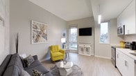 This single-floor space showcases a thoughtfully designed living area that balances modern aesthetics with functional comfort. The room features vaulted ceilings that amplify the sense of openness, complemented by light hardwood flooring that runs throughout and contributes to the overall bright atmosphere.

Natural light flows generously into the space through multiple windows, offering views of the surrounding outdoor environment. The neutral gray walls serve as a versatile backdrop, allowing the carefully chosen furnishings to stand out, most notably a vibrant yellow accent chair that adds a bold pop of color against the muted tones.

The living area is well-appointed with a gray sectional sofa, a geometric glass-top coffee table, and a plush area rug that defines the seating zone. A wall-mounted flat-screen television is positioned opposite the seating arrangement, flanked by a white console table with matching stools beneath. Decorative elements, including abstract wall art and carefully placed accessories, add character and visual interest throughout the space. The overall ambiance is clean, contemporary, and inviting.