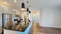 Located at 4541 Helm Dr in Virginia Beach, Virginia, this well-appointed single-floor residence offers a thoughtfully designed layout with modern finishes throughout. The kitchen serves as a centerpiece of the home, featuring a large island with striking dark granite countertops, a deep sink, stainless steel appliances including a refrigerator, microwave, and range, and crisp gray shaker-style cabinetry. Three bold black pendant lights hang above the island, adding a contemporary touch, while recessed lighting and subway tile backsplash complete the polished look. Warm hardwood-style flooring flows through the kitchen and main living areas, creating a cohesive and inviting atmosphere. A spacious living area with crown molding and a glass door leads to a private balcony with outdoor views, filling the space with natural light. The bedrooms are carpeted for comfort and offer generous windows that bring in abundant daylight. The primary bathroom features a dual-sink vanity with white cabinetry, dark hardware accents, a large mirror, and a built-in linen tower, with an adjacent view into the carpeted bedroom. A second full bathroom includes a tub-shower combination and wood-look flooring. A dedicated utility room houses a full-size washer and water heater, providing added convenience within the home.