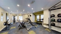 This fitness center is designed to provide a comprehensive workout experience within a single-level space. The room features a neutral color palette accented by olive-green wall details, creating a modern and energizing atmosphere. Recessed ceiling lights, complemented by natural light streaming through multiple windows dressed with roman shades, ensure the space remains bright and welcoming throughout the day.

The flooring consists of patterned carpet tiles in gray and gold tones, adding visual interest while providing a cushioned surface underfoot. The cardio section is equipped with upright exercise bikes, elliptical machines, and treadmills arranged to allow comfortable spacing between users. A flat-screen TV is mounted on the wall, providing entertainment during workouts.

The strength training area features a well-stocked dumbbell rack with a full range of free weights neatly organized on tiered shelving, accompanied by a padded workout bench. Additional cable and resistance machines are positioned along the perimeter, maximizing the use of available floor space. The overall layout is thoughtfully arranged to accommodate a variety of fitness routines efficiently.