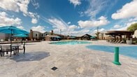This outdoor amenity space is designed as a resort-style recreational area, centered around a generously sized freeform swimming pool with crystal-clear blue water. The pool's organic, curved shape lends a relaxed, leisure-oriented aesthetic, and its surrounding travertine tile deck provides ample space for lounging and entertaining. Bright blue umbrellas and lounge chairs are positioned throughout the deck, offering shaded spots to unwind under the open sky.

A prominent covered pavilion serves as a central gathering point, featuring a peaked roofline with a rustic timber frame design, outdoor seating, and what appears to be a built-in bar or kitchen area — ideal for social gatherings and events. A wood pergola on the opposite side of the pool provides an additional shaded retreat with chaise lounges.

Adjacent to the pool area is a dedicated splash pad, surfaced in vibrant blue with colorful water play features, making it a family-friendly addition to the complex. The surrounding landscape includes manicured greenery, raised planting beds, and mature trees that frame the space.

The entire area is enclosed by iron fencing with views of the surrounding multi-story brick and siding residential buildings. The open-sky setting allows abundant natural light throughout the day, creating a bright and inviting outdoor environment suited for year-round enjoyment.