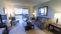 This single-floor space offers a comfortable and thoughtfully designed living environment. The living room serves as the centerpiece, bathed in natural light streaming through a large sliding glass door that opens onto a private balcony with a railing — offering a pleasant outdoor view of surrounding trees. The balcony extends the sense of space and provides a seamless indoor-outdoor connection.

Inside, the living room features plush neutral carpeting underfoot, creating a warm and inviting atmosphere. A striking blue accent wall adds a pop of color and visual depth to the otherwise soft, neutral palette of the room. The space is generously proportioned, comfortably accommodating a full sofa, accent chair, and a substantial coffee table without feeling crowded.

A decorative framed mirror on the accent wall enhances the sense of openness by reflecting light throughout the room. Soft ambient lighting from a floor lamp complements the natural light, creating a cozy atmosphere during the evening hours. The textured ceiling adds a subtle architectural detail overhead.

Overall, this space strikes a balance between functional design and comfortable living, with well-considered details that create a relaxed, inviting ambiance throughout.