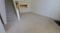 This two-floor space offers a practical and open layout designed for comfortable everyday living. The main level centers around a generously sized living area finished with neutral-toned wall-to-wall carpeting, creating a warm and cohesive atmosphere throughout. White walls and a clean, minimal aesthetic give the space a bright, open feel with ample room to arrange furnishings to suit a variety of needs.

A white-railed staircase positioned along one side of the living area provides access to the second floor, adding both functional connectivity and a subtle architectural detail to the main level. Adjacent to the living area, the kitchen features warm wood-toned cabinetry and is connected to the living space via a convenient pass-through window, promoting an open flow between the cooking and living areas. A refrigerator is visible within the kitchen, and the layout appears well-suited for efficient use of space.

The overall design reflects a straightforward and functional floor plan with neutral finishes throughout, making the space highly adaptable. The combination of carpeted living areas, the open kitchen connection, and the two-level layout provides a well-rounded and livable environment suitable for a range of occupants.