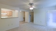 This single-floor space features an open and functional layout with a bright, neutral color palette of white and beige tones throughout. The main living area is finished with wall-to-wall carpet and is anchored by a ceiling fan with an integrated light fixture, providing both comfort and illumination. A textured ceiling adds subtle character to the space.

A pass-through window connects the living area to an adjacent kitchen or utility space, enhancing the open feel and allowing for easy interaction between rooms. Built-in shelving is visible in a recessed nook, offering practical storage and display options without occupying additional floor space. A built-in desk or countertop surface is also present in this area, adding versatile functionality.

Toward the rear of the space, a hallway leads to additional rooms, with a door at the end featuring a small window that allows natural light to filter through. The layout appears well-organized, with clear sightlines from the main living area through to the back of the unit. White walls and consistent flooring contribute to a clean, cohesive aesthetic throughout the space.
