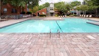 Located at 1300 Longcreek Dr in Columbia, South Carolina, this single-level outdoor amenity space centers around a generously sized swimming pool with clear, blue water. The pool ranges from shallow to moderate depth, with stainless steel entry rails and clearly marked depth indicators along the brick paver pool deck. The surrounding patio is finished with attractive, textured brick pavers that extend across the entire outdoor area, providing a cohesive and durable surface throughout. Numerous sling-style lounge chairs are arranged poolside, offering ample space for relaxation and sunbathing. A covered pavilion supported by brick columns provides a shaded retreat adjacent to the pool, featuring additional seating and overhead lighting. A picnic table is also available within the pavilion for outdoor dining. The space is framed by mature trees and well-maintained landscaping, including hedgerows and decorative plantings that add a sense of privacy and natural ambiance. The surrounding multi-story brick and siding residential buildings are visible in the background, complementing the community-oriented character of the space. Umbrellas and additional seating areas are scattered throughout, making this an inviting and functional outdoor gathering space suited for warm South Carolina days.