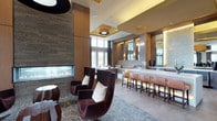 This stylish single-floor amenity space is designed with a contemporary aesthetic that blends warm wood tones, soft neutral finishes, and carefully curated furnishings to create an inviting, upscale atmosphere. The layout flows across multiple interconnected zones, each serving a distinct purpose while maintaining a cohesive design language throughout.

A central lounge area anchors the space, featuring plush, deep-toned velvet armchairs arranged around a sleek linear gas fireplace framed by a textured stone surround and warm wood paneling. Overhead, a bold drum pendant light with a gold-toned ceiling inset adds a striking focal point. Patterned carpet in soft grey tones with organic line motifs runs throughout, tying the areas together elegantly.

Adjacent to the lounge, a bar with a long white countertop and warm-toned bar seating provides a social gathering spot, enhanced by backlit mirrors and ambient lighting. A games area includes a full-size billiards table positioned near floor-to-ceiling windows that frame outdoor views and flood the space with natural light.

A flexible meeting and activity area features high-top tables, a game table, and a decorative built-in shelving unit with illuminated display niches. A utility area with light wood cabinetry and tile flooring completes the space with functional storage and prep capabilities.