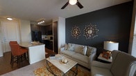 This single-floor residence showcases a well-designed open-concept layout that seamlessly connects the living area and kitchen. The living room draws immediate attention with a dramatic dark charcoal accent wall, serving as a striking backdrop for decorative starburst mirrors that add a touch of contemporary flair. A neutral-toned sofa with patterned throw pillows anchors the seating area, complemented by a metal-framed coffee table and a warm, patterned area rug that adds color and texture underfoot. A ceiling fan with an integrated light fixture overhead provides both comfort and illumination. Carpeted flooring runs throughout the living space, contributing to a cozy atmosphere. The adjoining kitchen features dark cabinetry paired with light granite-style countertops, black appliances, and track lighting above, creating a sleek, modern aesthetic. A kitchen peninsula provides a natural transition between the two spaces, enhancing the open flow of the layout. Overall, the space balances bold design choices with warm, inviting elements to create a cohesive and functional living environment.