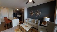 This single-floor residence showcases a well-designed open-concept layout that seamlessly connects the living area and kitchen. The living room draws immediate attention with a dramatic dark charcoal accent wall, serving as a striking backdrop for decorative starburst mirrors that add a touch of contemporary flair. A neutral-toned sofa with patterned throw pillows anchors the seating area, complemented by a metal-framed coffee table and a warm, patterned area rug that adds color and texture underfoot. A ceiling fan with an integrated light fixture overhead provides both comfort and illumination. Carpeted flooring runs throughout the living space, contributing to a cozy atmosphere. The adjoining kitchen features dark cabinetry paired with light granite-style countertops, black appliances, and track lighting above, creating a sleek, modern aesthetic. A kitchen peninsula provides a natural transition between the two spaces, enhancing the open flow of the layout. Overall, the space balances bold design choices with warm, inviting elements to create a cohesive and functional living environment.