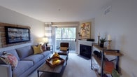 This single-floor residence offers a thoughtfully designed living space that balances comfort and style. The living room serves as the heart of the home, featuring plush carpeting, a classic fireplace with a clean white mantle, and a large sliding glass door that opens to a private balcony. The balcony view reveals lush palm trees, lending a warm, relaxed ambiance to the interior. Natural light fills the room through the generously sized glass door, complemented by striped curtains that add a soft, contemporary touch. The living area is appointed with modern furnishings, including a grey sofa accented with vibrant yellow throw pillows, a leather armchair, and a rustic wood coffee table. A bold cycling-themed artwork above the fireplace adds a creative focal point to the space. An open shelving console unit along one wall provides both display and storage opportunities, styled with candles and greenery. The overall aesthetic is warm and inviting, blending neutral tones with pops of gold and earthy accents, creating a cohesive and welcoming atmosphere throughout this single-level home.
