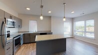 Located at 21 East St in North Attleboro, MA, this thoughtfully designed single-floor unit offers a clean, contemporary aesthetic throughout. The open-concept kitchen is a standout feature, showcasing granite countertops, gray shaker-style cabinetry with black hardware, and a full suite of stainless steel appliances including a refrigerator, range, microwave, and dishwasher. A large center island with matching granite provides additional prep and seating space, while stylish pendant lighting adds a finishing touch. The kitchen flows seamlessly into an open living area finished with light wood-look flooring and a ceiling fan, creating a bright and airy atmosphere. Natural light filters through multiple windows fitted with blinds throughout the unit. The bedrooms feature neutral-toned carpet, soft gray walls, and generous closet space with organized shelving. Two bathrooms serve the unit — one with a tub/shower combination and granite vanity, and another with grab bars and accessible fixtures, reflecting an inclusive design approach. Both bathrooms continue the granite countertop theme with white cabinetry and wood-look flooring. The overall design palette of soft neutrals, modern finishes, and practical layouts makes this a comfortable and functional living space.