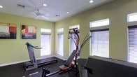 Located in Amarillo, Texas, this single-floor fitness space is designed to support a comprehensive workout experience. The exercise room is bright and functional, featuring large windows fitted with horizontal blinds that allow generous amounts of natural light to fill the space. High-placed transom windows add additional light while maintaining privacy. Recessed ceiling lights supplement the natural illumination, and a ceiling fan ensures comfortable airflow during workouts. The walls are finished in a soft, light green tone, creating an energizing yet calm atmosphere. The room is equipped with a variety of exercise machines, including a treadmill and a multi-function cable resistance training system, accommodating a range of fitness routines. The flooring is dark and durable, suitable for high-traffic fitness use. A piece of wall art adds a touch of personality to the space. Overall, the facility is clean, well-lit, and thoughtfully arranged to maximize usability and comfort for its occupants.