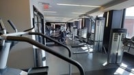 This fitness center offers a well-organized, single-floor layout designed to accommodate a comprehensive workout experience. The space features a sleek, contemporary aesthetic with dark accent walls contrasting against a light ceiling fitted with recessed linear lighting that illuminates the room evenly. Large windows along one side of the facility allow natural light to flood in, enhancing the open and energizing atmosphere.

The cardio section is prominently featured, housing a row of treadmills and elliptical machines that extend the length of the space, creating an efficient and accessible workout corridor. On the opposite side, a selection of Life Fitness strength training machines and cable systems are strategically arranged to allow for smooth traffic flow between equipment. Rubber flooring throughout provides both safety and durability underfoot.

Mirrored surfaces along portions of the walls add depth to the space while serving a functional purpose for form-checking during exercise. The overall ambiance is clean, professional, and well-maintained, reflecting a facility equipped to meet a broad range of fitness needs in a comfortable and modern environment.