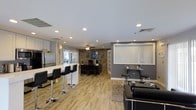 This single-floor space offers an open, versatile layout that seamlessly integrates multiple functional zones. The interior is finished with light wood-look flooring throughout, creating a cohesive and warm aesthetic across the entire area. Recessed lighting illuminates the space evenly, complemented by a ceiling fan that adds both style and airflow.

A kitchen bar area anchors one side of the space, featuring a white countertop and sleek black bar stools, alongside stainless steel appliances visible in the background. French doors provide access to an outdoor area, allowing natural light to filter in.

A dedicated workspace sits centrally, defined by an exposed brick accent wall that adds texture and character. A desk setup with a monitor makes this area suitable for professional use. Adjacent to this, vertical blinds frame a window on a gray accent wall, near a bistro-style seating arrangement ideal for casual meetings or relaxation.

The lounge area rounds out the space with comfortable seating and a decorative area rug. The combination of modern finishes, exposed brick, and flexible zones makes this space functional and visually engaging.