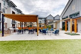 Located at 16200 Croft Drive in Charlotte, NC, this thoughtfully designed outdoor amenity space serves as a central courtyard hub for community gathering and recreation. The grounds-level space blends modern farmhouse architectural elements — including board-and-batten siding, metal roofing accents, and warm wood pergola structures — with a highly functional outdoor layout.

The courtyard is anchored by multiple distinct zones. A generously sized open lawn area of lush green turf provides space for recreation and lawn games, including a cornhole board. Adjacent to the lawn, a covered pavilion equipped with a wall-mounted television and comfortable blue lounge seating offers a sheltered retreat for outdoor entertainment.

A large paved patio serves as the social heart of the space, featuring multiple seating arrangements with cushioned chairs gathered around low tables, ideal for casual conversation. A striking horizontal wood privacy screen creates a defined backdrop for one of the lounge areas, adding warmth and visual interest. Overhead string lights add an inviting ambiance for evening gatherings.

A dedicated outdoor kitchen and dining area, positioned beneath a wood pergola, includes a built-in grill and ample table seating for group dining. Colorful planted flower beds and manicured landscaping are woven throughout, softening the hardscape and enhancing the overall aesthetic. Large sliding glass doors connect the outdoor space to an adjacent interior amenity area, ensuring seamless indoor-outdoor flow.