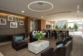 This elegantly designed single-floor amenity lounge offers a seamless blend of contemporary style and comfortable functionality. The space is anchored by a striking bar area complete with a dark granite countertop, rich wood cabinetry, bar-height seating, a mirrored backsplash with stone tile accents, and a built-in beverage refrigerator — ideal for entertaining and social gatherings.

Adjacent to the bar, a spacious lounge area features plush leather seating arrangements, a statement circular pendant light fixture, and recessed cove lighting that adds warmth to the refined neutral palette. Framed artwork punctuates the walls, lending a curated, gallery-like atmosphere.

A welcoming entry corridor introduces the space with warm wood-paneled walls, tile flooring, and a cozy built-in bench seating nook, setting an inviting tone from the moment of arrival.

Throughout the lounge, floor-to-ceiling windows flood the interior with natural light and frame sweeping panoramic views of the surrounding cityscape and greenery beyond. Sheer drapery softens the light while maintaining an open, airy feel. The flooring transitions thoughtfully between light tile and textured carpet, defining each zone while maintaining a cohesive, upscale aesthetic. Together, these elements create a polished and inviting communal environment.
