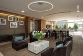 This elegantly designed single-floor amenity lounge offers a seamless blend of contemporary style and comfortable functionality. The space is anchored by a striking bar area complete with a dark granite countertop, rich wood cabinetry, bar-height seating, a mirrored backsplash with stone tile accents, and a built-in beverage refrigerator — ideal for entertaining and social gatherings.

Adjacent to the bar, a spacious lounge area features plush leather seating arrangements, a statement circular pendant light fixture, and recessed cove lighting that adds warmth to the refined neutral palette. Framed artwork punctuates the walls, lending a curated, gallery-like atmosphere.

A welcoming entry corridor introduces the space with warm wood-paneled walls, tile flooring, and a cozy built-in bench seating nook, setting an inviting tone from the moment of arrival.

Throughout the lounge, floor-to-ceiling windows flood the interior with natural light and frame sweeping panoramic views of the surrounding cityscape and greenery beyond. Sheer drapery softens the light while maintaining an open, airy feel. The flooring transitions thoughtfully between light tile and textured carpet, defining each zone while maintaining a cohesive, upscale aesthetic. Together, these elements create a polished and inviting communal environment.