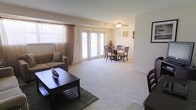 This single-floor residential space offers an open, airy layout that seamlessly connects the living and dining areas into one cohesive environment. The living room is carpeted in plush, light-toned flooring that extends throughout, creating a warm and inviting atmosphere. Large windows dressed with sheer curtains allow abundant natural light to filter in, while a set of full-length glass patio doors at the far end of the room enhances the brightness and provides a direct connection to the outdoors. A ceiling fan with a light fixture serves as a practical overhead feature, ensuring comfort throughout the space. The living area comfortably accommodates a sofa with decorative pillows and a dark wood coffee table set atop a textured area rug, providing a cozy setting for relaxation. Adjacent to the living room, the dining area features a round table surrounded by wooden chairs, positioned conveniently near the patio doors to take advantage of the natural light. The walls are finished in a neutral, soft tone that complements the overall calm and clean aesthetic of the space. The layout is efficient and well-proportioned, making it both functional and welcoming for everyday living.