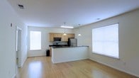 This single-floor space is designed around an open-concept layout that seamlessly integrates the kitchen and living areas into one cohesive environment. The kitchen is well-appointed with light maple cabinetry, dark countertops, and a full suite of stainless steel appliances, including a refrigerator, range, and built-in microwave. A peninsula counter serves as a functional divider between the kitchen and the open living area, offering additional workspace and the potential for casual seating. Pendant lights hang above the peninsula, providing focused task lighting, while a linear ceiling fixture illuminates the kitchen workspace. The living area benefits from light hardwood flooring that runs throughout, contributing warmth and continuity across the space. Multiple windows fitted with horizontal blinds allow generous amounts of natural light to fill the interior, creating a bright and airy atmosphere. The neutral wall tones complement the overall clean and modern aesthetic of the space. The layout is efficient and practical, making the most of the available footprint while maintaining an open, uncluttered feel throughout.