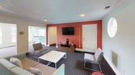 This single-floor community lounge is thoughtfully designed to offer a variety of functional zones within one cohesive space. Upon entry, a welcoming corridor with dark carpet flooring and white trim leads into the main gathering area, with a narrow console table providing a tasteful transitional accent. A glass-paned door opens directly to an adjacent fitness center, offering convenient connectivity between amenity spaces.

The lounge is anchored by a spacious seating area featuring a neutral sofa accented with colorful throw pillows, a clean-lined white coffee table, and an assortment of accent chairs arranged around a wall-mounted flat-screen television set against a bold terracotta-red feature wall. This warm accent wall creates a striking focal point and adds character to the room.

Adjacent to the main seating area, a lighter zone finished in soft aqua-toned walls offers a more relaxed retreat, complete with a built-in ledge, additional seating, and a distinctive circular window that floods the space with natural light alongside a standard double-hung window. A separate dining nook features a sleek round bistro table with contemporary chrome-legged bar stools, positioned near a glass door with views to an outdoor area. Crown molding and textured ceilings lend a polished, finished quality throughout the space.