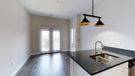 Highland Station presents a thoughtfully designed single-floor residence that blends modern aesthetics with everyday functionality. The open-concept living and kitchen area serves as the heart of the home, featuring rich dark hardwood-style flooring, neutral warm-toned walls, and a set of elegant French doors that flood the space with natural light and provide access to an outdoor balcony. Stylish black pendant lights hang above the kitchen, which is equipped with stainless steel appliances, including a range, over-the-range microwave, and full-size refrigerator. Crisp white cabinetry, dark granite countertops, and a classic white subway tile backsplash complete the kitchen's polished look. A convenient in-unit laundry area is tucked just off the kitchen. The bedroom is a calm retreat, carpeted for comfort, with large windows fitted with blinds that allow for ample natural light and a ceiling fan for year-round comfort. The bathroom features a full soaking tub and shower combination, elegantly lined with large-format marble-look tile, paired with brushed nickel fixtures. An adjacent walk-in closet with built-in shelving adds practical storage. Throughout the residence, clean lines, contemporary finishes, and a cohesive neutral palette create a bright, inviting atmosphere.