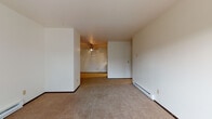 This single-floor unit offers a functional and straightforward layout spread across several connected spaces. The living area is generously sized, featuring neutral-toned carpet underfoot and clean white walls throughout. A ceiling fan provides air circulation, while a baseboard heating system runs along the perimeter walls, ensuring year-round comfort. A sliding glass door opens to a private balcony that overlooks mature trees, bringing natural light into the space and offering a pleasant outdoor connection. Built-in shelving flanks the balcony entrance, adding practical storage to the living area.

The galley-style kitchen is outfitted with warm oak cabinetry providing ample upper and lower storage, white countertops, a stainless steel sink, and black appliances including a refrigerator and range. Tile flooring adds durability to this workspace.

The bedroom is a well-proportioned room with carpeting, a window offering natural light and tree views, and a wide closet with bi-fold doors and a full-length hanging rod. Baseboard heaters line the walls here as well.

The bathroom features a full-sized soaking tub and shower with white tiled surround, complemented by tile flooring. Overall, the unit presents a clean, neutral palette throughout.