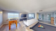 This single-floor entertainment space is thoughtfully designed to combine relaxed lounge areas with recreational amenities, all set against stunning waterfront views. The open layout flows seamlessly across light hardwood-style flooring, with distinct zones defined by area rugs and subtle architectural transitions rather than walls.

The main gathering area centers around a white sectional sofa and a low-profile coffee table, anchored by a wall-mounted flat-screen TV positioned above a sleek navy and gold credenza. Large windows frame panoramic water views, flooding the space with natural light and creating a bright, airy atmosphere throughout.

Adjacent to the lounge, a rustic wood-framed pool table serves as a focal point of the games area, complemented by a foosball table nearby. Wall sconces provide warm accent lighting in the game zones, while overhead flush-mount fixtures illuminate the lounge areas evenly.

A secondary seating alcove connects to the main space, furnished with cream accent chairs, a long coffee table, and additional waterfront-facing windows. The neutral white walls and textured ceilings maintain a clean, contemporary aesthetic, while the consistent flooring and open sightlines unify the entire space into one cohesive, inviting environment.