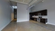 This single-floor space showcases a striking industrial-modern aesthetic defined by soaring ceilings with exposed wooden planks and visible ductwork. The open-concept layout seamlessly connects the main living area to the kitchen, creating a spacious and airy environment. Polished concrete floors run throughout, adding a sleek, contemporary foundation that complements the industrial character of the space. The kitchen is well-appointed with dark espresso cabinetry, stainless steel appliances including a refrigerator, microwave, range, and dishwasher, as well as a clean white countertop with an undermount sink. Track lighting above the kitchen provides focused illumination, while pendant lighting extends into the open living area. The tall, uninterrupted accent wall serves as a dramatic architectural feature, enhancing the sense of vertical space. A hallway visible in the background leads to additional areas of the unit, offering functional separation within the open design. Overall, the space balances raw industrial elements with modern finishes, creating a cohesive and contemporary atmosphere.