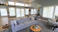 This single-floor space makes a strong impression with its vaulted ceilings and generous use of natural light. Clerestory windows positioned high along the roofline, combined with multiple large windows along the lower walls, flood the interior with daylight and create an airy, open atmosphere throughout.

The main living area is anchored by a substantial gray sofa dressed with patterned throw pillows, paired with a natural wood and metal coffee table. The layout flows effortlessly into a defined dining area, where a round wood table is surrounded by sleek black metal chairs beneath an industrial-style orb chandelier, adding a stylish focal point overhead.

The interior palette features cool gray walls complemented by bright white trim and window casings, lending the space a clean, contemporary feel. Patterned carpet in a neutral tone grounds the living zone, while the consistent design language ties both areas together cohesively.

White plantation shutters throughout offer light control while maintaining the space's polished aesthetic. A decorative wall clock and subtle accent lighting round out the thoughtfully curated design, resulting in a welcoming and functional environment.