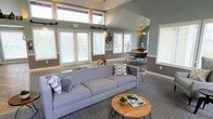 This single-floor space makes a strong impression with its vaulted ceilings and generous use of natural light. Clerestory windows positioned high along the roofline, combined with multiple large windows along the lower walls, flood the interior with daylight and create an airy, open atmosphere throughout.

The main living area is anchored by a substantial gray sofa dressed with patterned throw pillows, paired with a natural wood and metal coffee table. The layout flows effortlessly into a defined dining area, where a round wood table is surrounded by sleek black metal chairs beneath an industrial-style orb chandelier, adding a stylish focal point overhead.

The interior palette features cool gray walls complemented by bright white trim and window casings, lending the space a clean, contemporary feel. Patterned carpet in a neutral tone grounds the living zone, while the consistent design language ties both areas together cohesively.

White plantation shutters throughout offer light control while maintaining the space's polished aesthetic. A decorative wall clock and subtle accent lighting round out the thoughtfully curated design, resulting in a welcoming and functional environment.