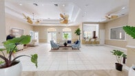 Located at 5500 NW 61st St in Coconut Creek, Florida, this single-story community clubhouse offers a generously proportioned open-plan interior designed for social gatherings and relaxation. The space is defined by its refined architectural details, including elevated tray ceilings with crown molding and recessed lighting that lend a polished, upscale atmosphere throughout.

The main gathering area is anchored by large-format tile flooring in neutral tones, creating a bright and cohesive look. Multiple seating arrangements — featuring crisp white leather sofas, tufted navy accent chairs, and patterned blue-and-white armchairs — provide comfortable, stylish areas for residents to congregate. Lush tropical potted plants are strategically placed throughout, reinforcing the Florida aesthetic.

A dedicated lounge section features a bar-height counter with seating, ideal for casual socializing. The grand entrance foyer welcomes visitors with decorative tile flooring, a statement pendant light fixture, and a set of glass-paned French doors flanked by arched transom windows.

Multiple sets of French doors with arched transoms line the perimeter, flooding the interior with natural light and offering direct views and access to an outdoor pool and patio area. Tropical-style ceiling fans provide both functional comfort and a resort-inspired design touch, completing the welcoming, sun-soaked ambiance of this Florida clubhouse.
