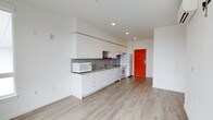 This single-floor unit offers a clean, contemporary design with an open-plan layout that maximizes usable space. The kitchen is the focal point of the main living area, featuring white flat-front cabinetry that extends to the ceiling for generous storage, complemented by a grey mosaic tile backsplash that adds texture to the otherwise minimal aesthetic. The countertops run along a full wall, providing ample food preparation space, while the kitchen is equipped with essential appliances including a refrigerator, electric range, built-in oven, and microwave. A stainless steel sink with a modern faucet is integrated seamlessly into the counter. Recessed ceiling lighting ensures bright, even illumination throughout the space. The flooring throughout is light-toned hardwood-style planks, contributing to a warm yet modern ambiance. A striking red entry door serves as a bold architectural accent, providing a vivid contrast against the predominantly white walls and neutral flooring. The open layout between the kitchen and living area creates a sense of spaciousness and flexibility, making the space highly functional for everyday living.