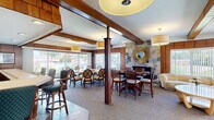 This well-appointed single-floor community room offers a comfortable and functional gathering space characterized by rich wood-paneled walls, exposed wooden beams, and warm pendant lighting throughout. The main lounge area features multiple seating arrangements, including a large round dining table surrounded by cushioned oval-back chairs positioned beside expansive sliding glass windows that look out onto lush greenery and an outdoor pool area, filling the room with natural light. A striking natural stone accent wall serves as a focal point, adorned with a decorative sunburst mirror and framing a fireplace, adding a classic, cozy ambiance. Bar-height seating along a counter provides additional casual gathering space, while a smaller private dining room with French doors and a round table offers a more intimate setting. The fully equipped kitchen features stainless steel appliances, including a refrigerator, range, and microwave, with rich wood cabinetry and a wraparound counter with bar stool seating, accented by colorful slate tile flooring. Throughout the space, carpet and tile flooring complement the warm wood tones, creating a cohesive, welcoming environment well-suited for social gatherings and community events.