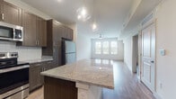 This thoughtfully designed single-floor residence offers a seamless open-concept layout that connects the kitchen and living area into one cohesive, light-filled space. The kitchen is a standout feature, showcasing rich dark wood cabinetry with sleek hardware, granite countertops, a white subway tile backsplash, and a full suite of stainless steel appliances including a refrigerator, range, and built-in microwave. A central island with a granite surface and elegant pendant lighting serves as both a functional workspace and a visual anchor for the space. Warm hardwood-style flooring flows consistently throughout the entire unit, lending a unified and contemporary feel. The living area is generously proportioned, benefiting from large windows fitted with horizontal blinds that allow abundant natural light to fill the room. A ceiling fan provides both comfort and a finishing touch to the open living space. The bedroom is spacious, featuring multiple windows, a ceiling fan, and access to a walk-in closet. The bathroom is cleanly appointed with a dark wood vanity and coordinating flooring. Throughout the space, recessed lighting and crisp white walls create a bright, modern ambiance that complements the warm tones of the cabinetry and flooring.