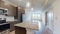 This thoughtfully designed single-floor residence offers a seamless open-concept layout that connects the kitchen and living area into one cohesive, light-filled space. The kitchen is a standout feature, showcasing rich dark wood cabinetry with sleek hardware, granite countertops, a white subway tile backsplash, and a full suite of stainless steel appliances including a refrigerator, range, and built-in microwave. A central island with a granite surface and elegant pendant lighting serves as both a functional workspace and a visual anchor for the space. Warm hardwood-style flooring flows consistently throughout the entire unit, lending a unified and contemporary feel. The living area is generously proportioned, benefiting from large windows fitted with horizontal blinds that allow abundant natural light to fill the room. A ceiling fan provides both comfort and a finishing touch to the open living space. The bedroom is spacious, featuring multiple windows, a ceiling fan, and access to a walk-in closet. The bathroom is cleanly appointed with a dark wood vanity and coordinating flooring. Throughout the space, recessed lighting and crisp white walls create a bright, modern ambiance that complements the warm tones of the cabinetry and flooring.