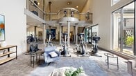 Located at 6200 Rime Village Dr NW in Huntsville, Alabama, this two-story fitness center offers a generously proportioned and thoughtfully designed space for health and wellness. The main floor commands attention with soaring ceilings, a row of stately decorative columns, and large windows that flood the area with natural light and outdoor views. The open layout accommodates a wide array of cardio equipment, including treadmills, ellipticals, and stationary bikes, all arranged to maximize space and usability. A comfortable seating area with leather chairs and a casual bar-height table with stools provides a welcoming spot to relax or socialize. A shiplap accent wall and a fireplace add warmth and character to the lower level. A curved staircase with sleek metal railings leads to the upper floor, where a mezzanine-level exercise area overlooks the main floor below. The second level features additional cardio machines, including recumbent bikes, positioned beside arched windows that frame lush exterior greenery. Wall-mounted televisions are present throughout both levels, enhancing the workout experience. Carpet tile flooring in a modern geometric pattern runs throughout, tying the two floors together with a cohesive, contemporary aesthetic.