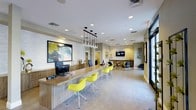 This single-floor commercial space offers a polished and contemporary environment designed to welcome and accommodate visitors with both style and functionality. The reception area anchors the space, featuring a long, warm-toned wood counter that provides ample workspace, complemented by bold yellow accent chairs that add a lively pop of color against the neutral palette. Overhead, a striking cluster of pendant lights with geometric glass elements serves as a focal point, while recessed lighting throughout ensures the space remains bright and inviting. The walls feature a mix of textured stone cladding and smooth painted surfaces, adding depth and visual interest to the overall design. A large piece of abstract wall art introduces vibrant color and artistic character near the reception desk. Toward the rear, a comfortable lounge area offers additional seating in muted, earthy tones, centered around a wall-mounted display screen — ideal for presentations or informational content. Large windows along the perimeter allow natural light to filter in, enhancing the open and airy atmosphere. The light-toned flooring ties the entire space together, creating a cohesive, professional, and approachable environment throughout.