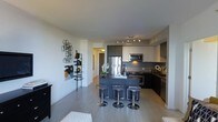 This single-floor space showcases a contemporary open-concept layout that seamlessly integrates the kitchen and living areas. The kitchen is a standout feature, equipped with stainless steel appliances, including a full-sized refrigerator and built-in microwave. Two-toned cabinetry — combining dark lower units with light upper cabinets — adds a sophisticated, modern aesthetic, while track lighting overhead ensures the workspace is well-illuminated.

A dark-finished central island serves as both a functional prep area and a social hub, accompanied by three adjustable chrome bar stools that invite casual dining or conversation. The countertops complement the cabinetry's contrast, reinforcing the space's cohesive design language.

Light-toned hardwood flooring runs throughout, contributing to a bright and airy atmosphere. The living area is visible in the foreground, hinting at a generous, flowing floor plan. A decorative geometric wall shelf adds a stylish, curated touch to the living space. Natural light appears to supplement the interior lighting, enhancing the overall sense of openness and warmth throughout the unit.