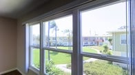 This single-story space offers a bright and inviting atmosphere, highlighted by expansive multi-pane windows that serve as a defining architectural feature. The large windows frame a picturesque outdoor scene, flooding the interior with abundant natural light and providing sweeping views of a well-maintained green courtyard below. Palm trees and neatly landscaped tropical plantings are visible just beyond the glass, evoking a warm, Southern California-style ambiance. The surrounding exterior reveals a low-rise residential community with classic mid-century architectural styling, characterized by clean lines, neutral tones, and manicured grounds with pathways winding through the greenery. The white-framed windows are thoughtfully designed to maximize the connection between the interior and the vibrant outdoor environment, making the space feel open, airy, and seamlessly integrated with its surroundings. The overall character of the space is defined by its excellent natural light, pleasant outdoor vistas, and a relaxed yet polished aesthetic that reflects the sunny, laid-back character of its locale. Whether used as a living area or gathering space, the visual relationship with the lush exterior landscape remains a central and standout element of the experience.
