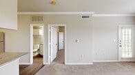 This 1-bedroom, 1-bathroom residence offers a thoughtfully organized layout across a single floor, encompassing seven distinct areas including a living room, kitchen, bedroom, walk-in closet, balcony, bathroom, and utility room. The living room is finished with plush carpeting, crown molding, and neutral warm-toned walls, creating a welcoming and versatile space. It connects seamlessly to the kitchen, which features white cabinetry, a subway tile backsplash, stainless steel appliances including a refrigerator and range, and durable hardwood-style flooring. The bedroom is a comfortable retreat with carpeted floors, large windows fitted with horizontal blinds that allow for ample natural light and outdoor views, and direct access to a spacious walk-in closet equipped with wire shelving and hanging rods on multiple walls. A built-in shelving nook with a desk surface adds a practical workspace element. The bathroom includes a full soaking tub and shower combination with decorative tile accents in neutral tones. Additional highlights include a private balcony for outdoor enjoyment and a dedicated utility room. Throughout the residence, crown molding, white panel doors, and consistent neutral finishes contribute to a clean, cohesive aesthetic.