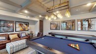 This well-appointed game and lounge space is designed for both recreation and relaxation, offering a thoughtfully curated environment on a single floor. The centerpiece of the room is a full-size billiards table with a bold blue felt surface, positioned beneath an eye-catching industrial-style chandelier fitted with amber globe pendants and brass accents. A shuffleboard table runs alongside, providing additional entertainment options for guests.

The coffered ceiling with exposed beam detailing adds architectural depth and visual interest overhead, while wood-look tile flooring throughout ties the space together with a warm, contemporary aesthetic. A wall-mounted flat-screen television makes the room equally suited for viewing as it is for gaming.

A cozy lounge nook features rich brown leather armchairs accented with colorful throw pillows, a round side table, and a floor lamp — creating an inviting retreat within the larger space. Framed industrial-themed artwork adorns the walls, contributing to the room's modern-rustic character.

Large sliding glass doors and expansive windows flood the space with natural light and offer views of the surrounding outdoor area, seamlessly connecting the interior to the exterior. A small bistro-style dining nook with draped curtains sits adjacent to the windows, providing an intimate spot for casual conversation or a quiet moment away from the games.