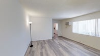 This single-floor space offers a clean and functional layout designed for comfortable everyday living. The main living area is generously open, finished with light gray wood-look laminate flooring that runs throughout, lending a cohesive and modern feel to the interior. Walls are painted in a crisp, neutral white, maximizing the brightness of the space and creating a welcoming atmosphere. A textured ceiling adds a classic touch overhead. Natural light filters in through a large window fitted with vertical blinds, contributing to the airy ambiance of the living area. A wall-mounted air conditioning unit ensures year-round climate comfort. The living area flows naturally toward a connecting hallway that leads to the kitchen, which is visible in the background and features white cabinetry and a bright interior. The straightforward, open-plan design makes the space feel practical and adaptable, suitable for a variety of furnishing arrangements. The overall aesthetic is simple and well-maintained, with clean lines and a neutral palette that provides a versatile canvas for personalization.