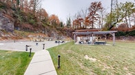 Located at 313 Creek Dr in Wayne, PA, The Yard is a thoughtfully designed outdoor gathering space that makes striking use of its natural surroundings. The space is dramatically framed by exposed stone rock faces and wooded hillsides, creating a distinctive and immersive natural backdrop that sets it apart from typical outdoor amenity areas.

A concrete walkway guides visitors through well-maintained green lawns and past large natural boulders that are intentionally incorporated into the landscape design. The centerpiece of the gravel activity area is a circular fire pit surrounded by Adirondack chairs, providing an inviting spot for relaxed social gatherings. Pathway bollard lighting is distributed throughout, ensuring the space remains functional and atmospheric into the evening hours.

A prominent covered pavilion, branded as "The Yard," anchors one side of the space and features a timber-frame structure with both open-air dining tables and lounge seating. Adjacent to the pavilion, a spacious gravel surface serves as a dedicated lawn games area, equipped for activities such as cornhole. The combination of rugged rock formations, lush turf, and curated amenities creates a versatile outdoor environment well-suited for both quiet relaxation and community gatherings.